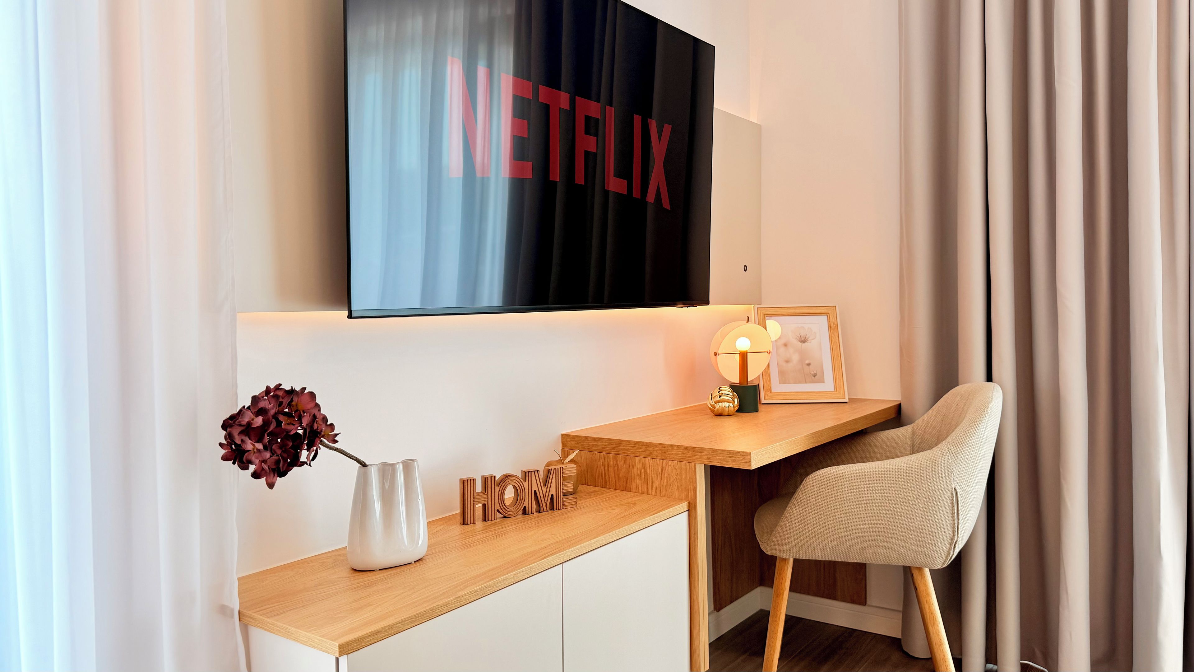 Habitación moderna con un escritorio de madera, una silla, un televisor montado en la pared mostrando 'NETFLIX', un jarrón con flores y un cuadro enmarcado.