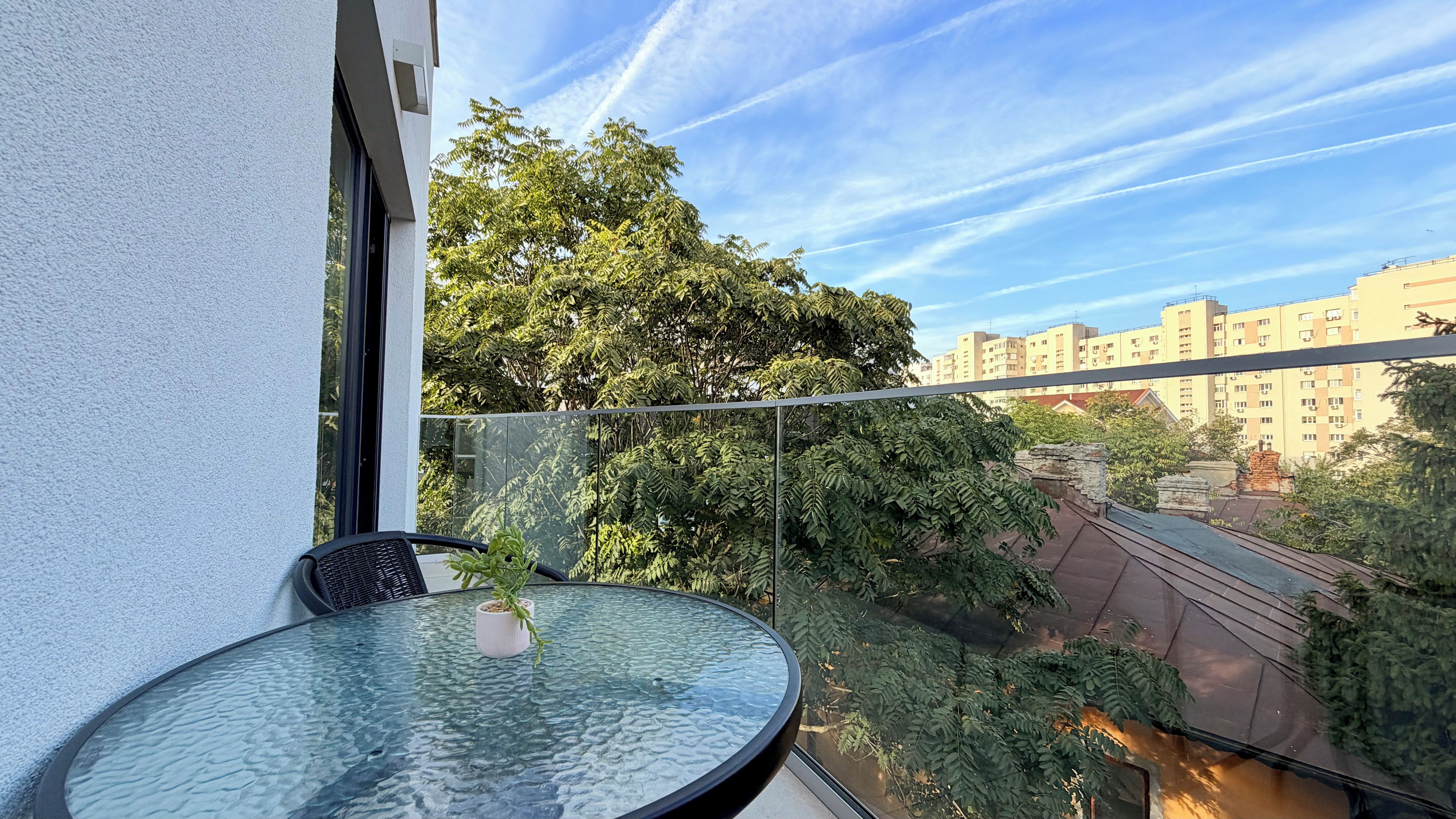 A balcony with a glass table and chair, overlooking a cityscape with buildings and trees.