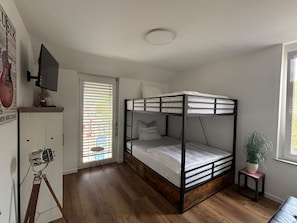 A compact room with a bunk bed, a small table with a plant, and a window with blinds. - New, Comfortable Vacation Rental for Families and Friends – 3 Minutes from the Beach (Bodman-Ludwigshafen)