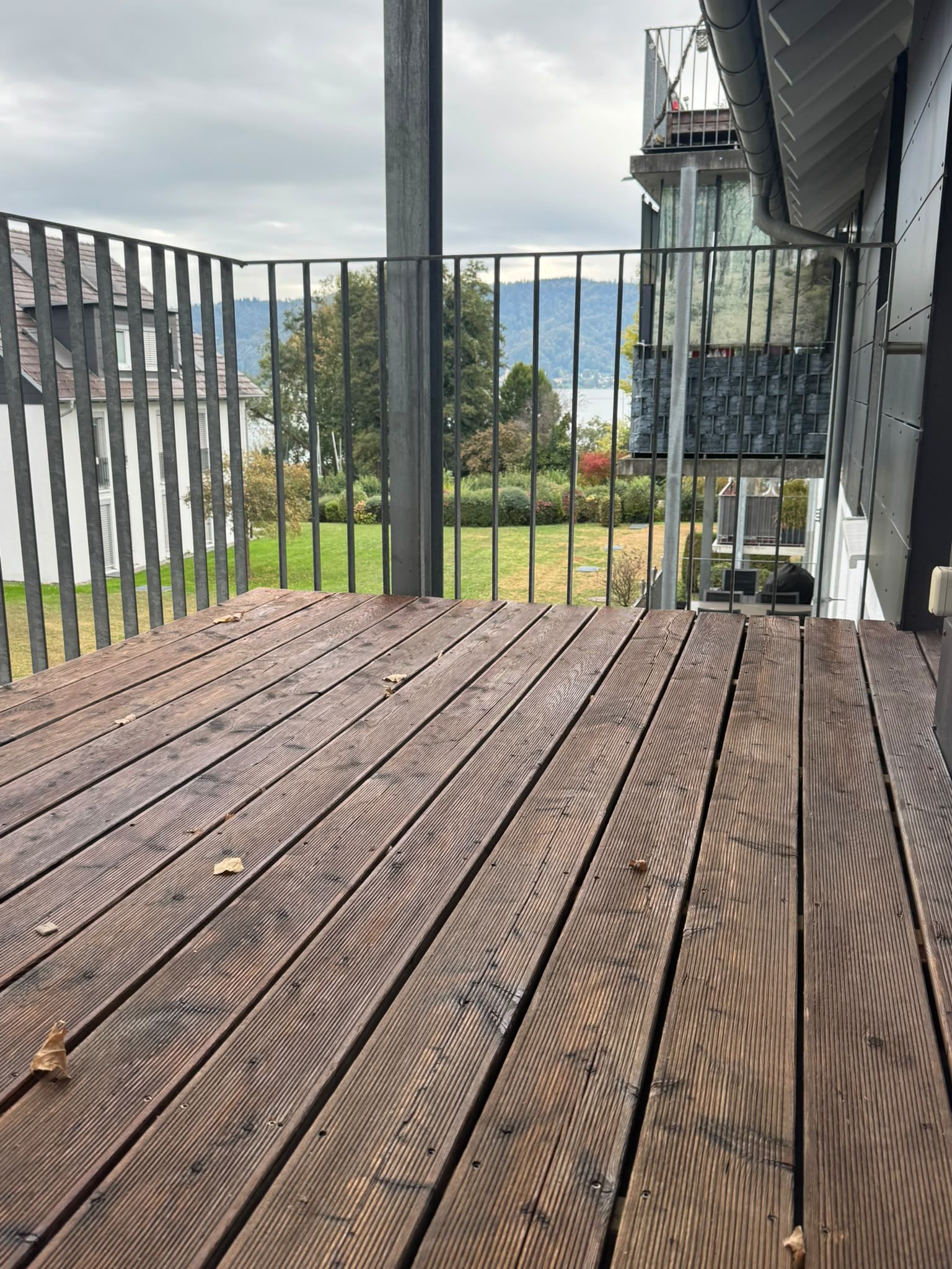 Une terrasse en bois avec une rambarde en métal, donnant sur un espace paysager agrémenté d’arbres et sur un bâtiment situé au loin.