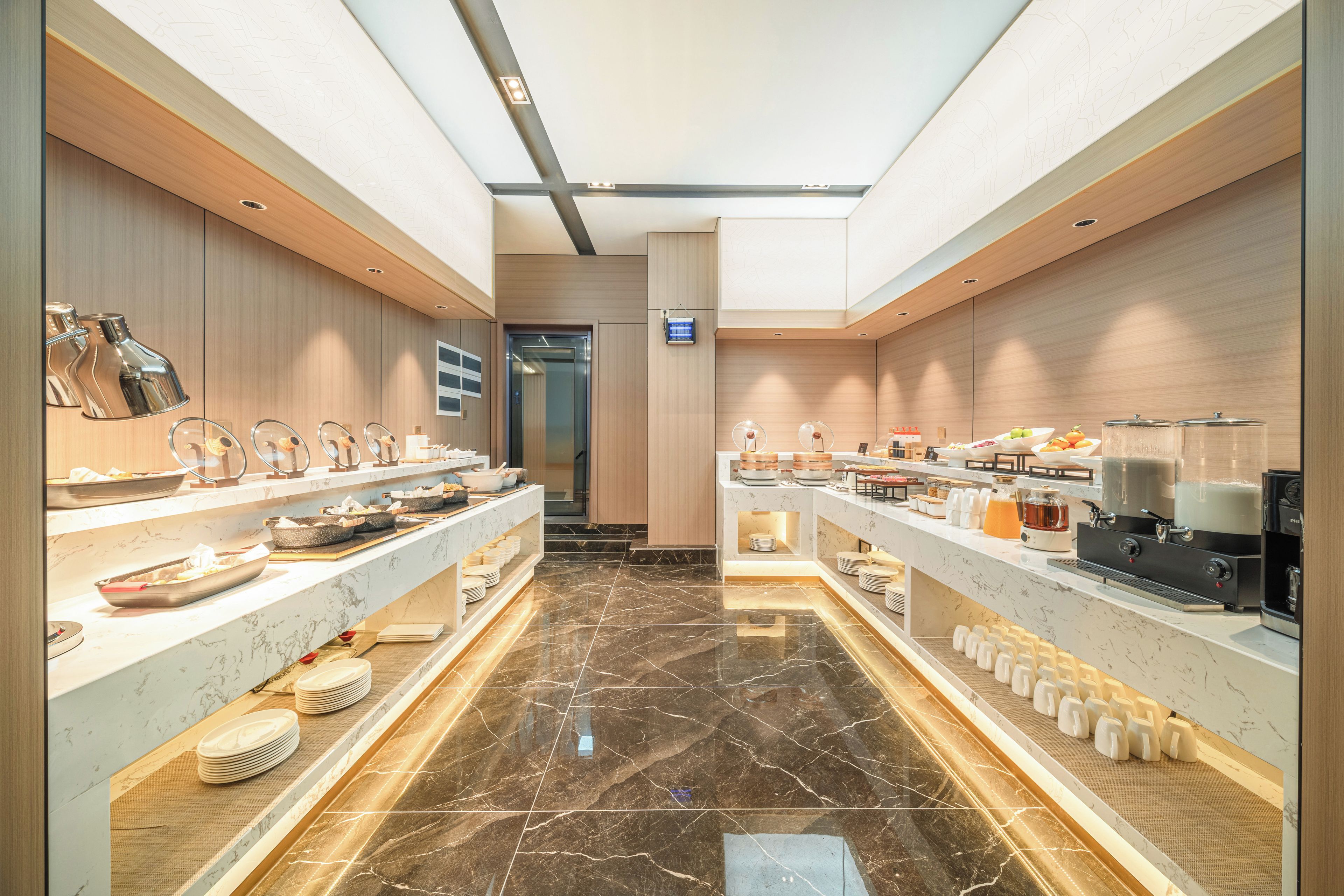 Un lobby moderno de hotel con suelo de mármol, un ascensor central y una zona de buffet con diversas opciones de comida y una estación de café.