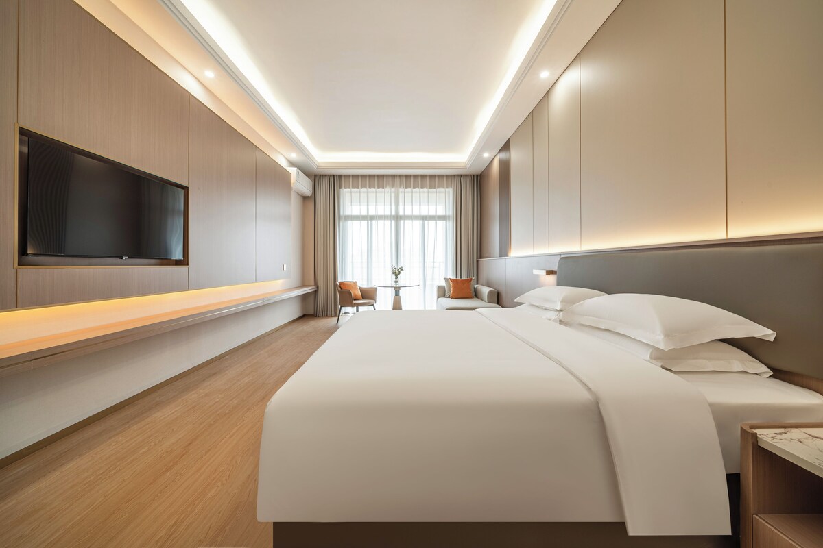 A hotel room with a large bed, a flat-screen TV mounted on the wall, a desk with a chair, and a window with curtains.