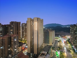 Un edificio alto con varios pisos, una entrada destacada y vistas de un paisaje urbano con otros edificios y una cadena montañosa distante.