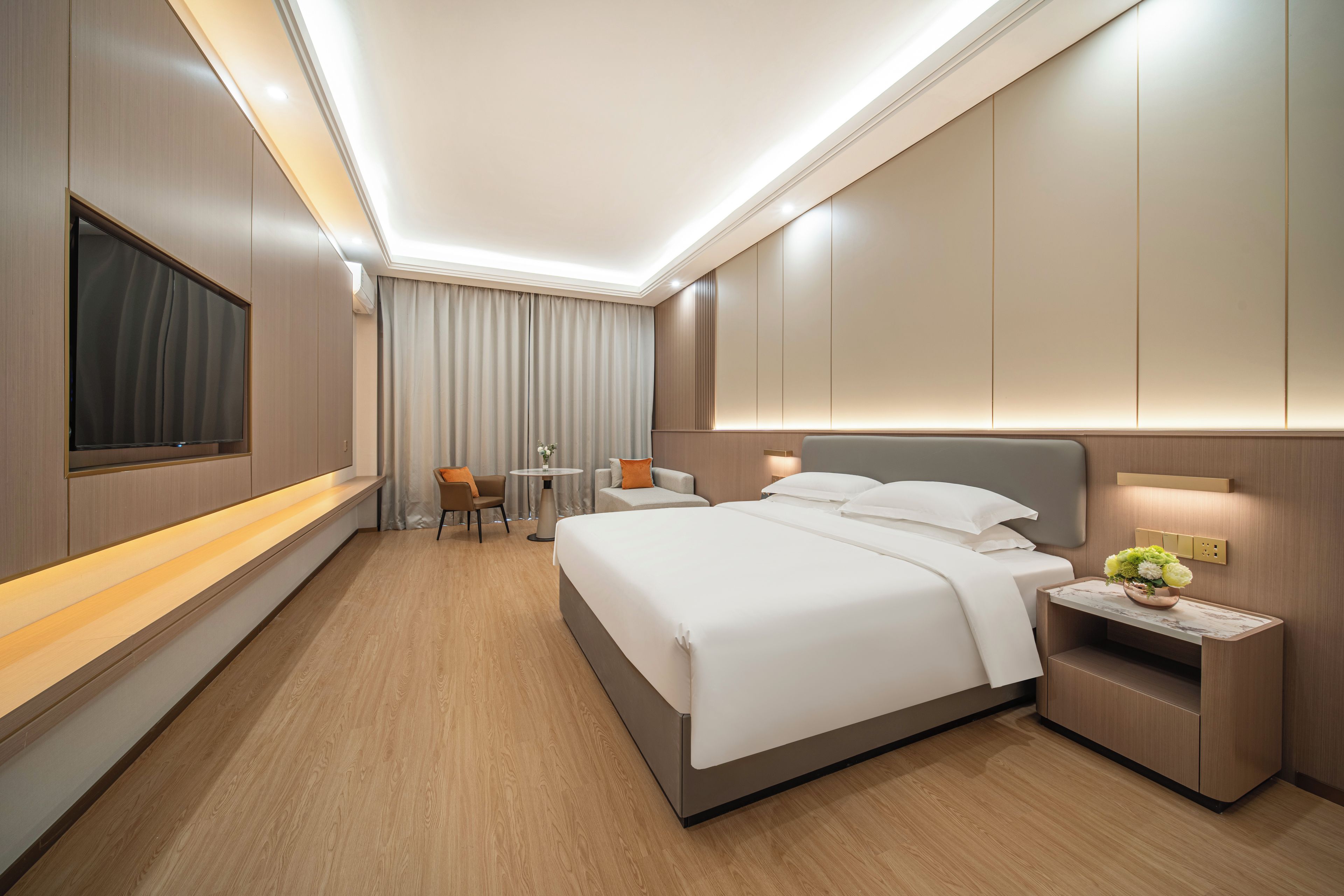 A modern hotel room with a large bed, bedside tables, a seating area with chairs, and a TV mounted on the wall.