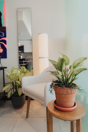 Un salon moderne avec un fauteuil blanc, une plante en pot posée sur un tabouret en bois, et une cuisine en arrière-plan.