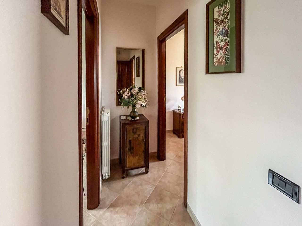 Ein Flur mit einem Heizkörper, einer Holzeinrichtung mit einer Blumenvase und einem gerahmten Kunstwerk an der Wand.