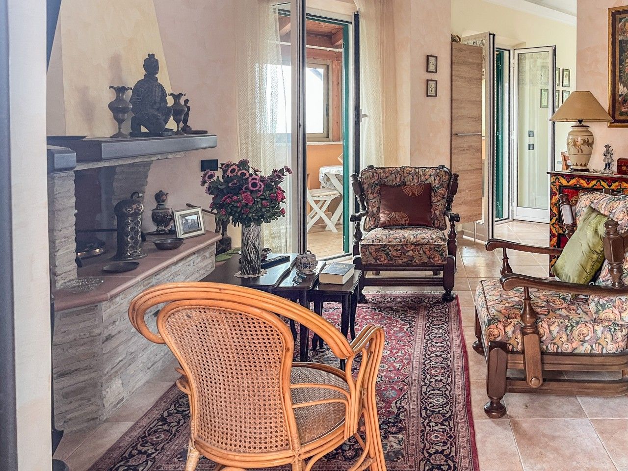 A cozy living room with a fireplace, a rattan chair, a floral sofa, and a dining area with a table and chairs.