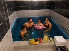 Una familia en un jacuzzi pequeño y con azulejos, acompañada de una bandeja de frutas y bebidas.