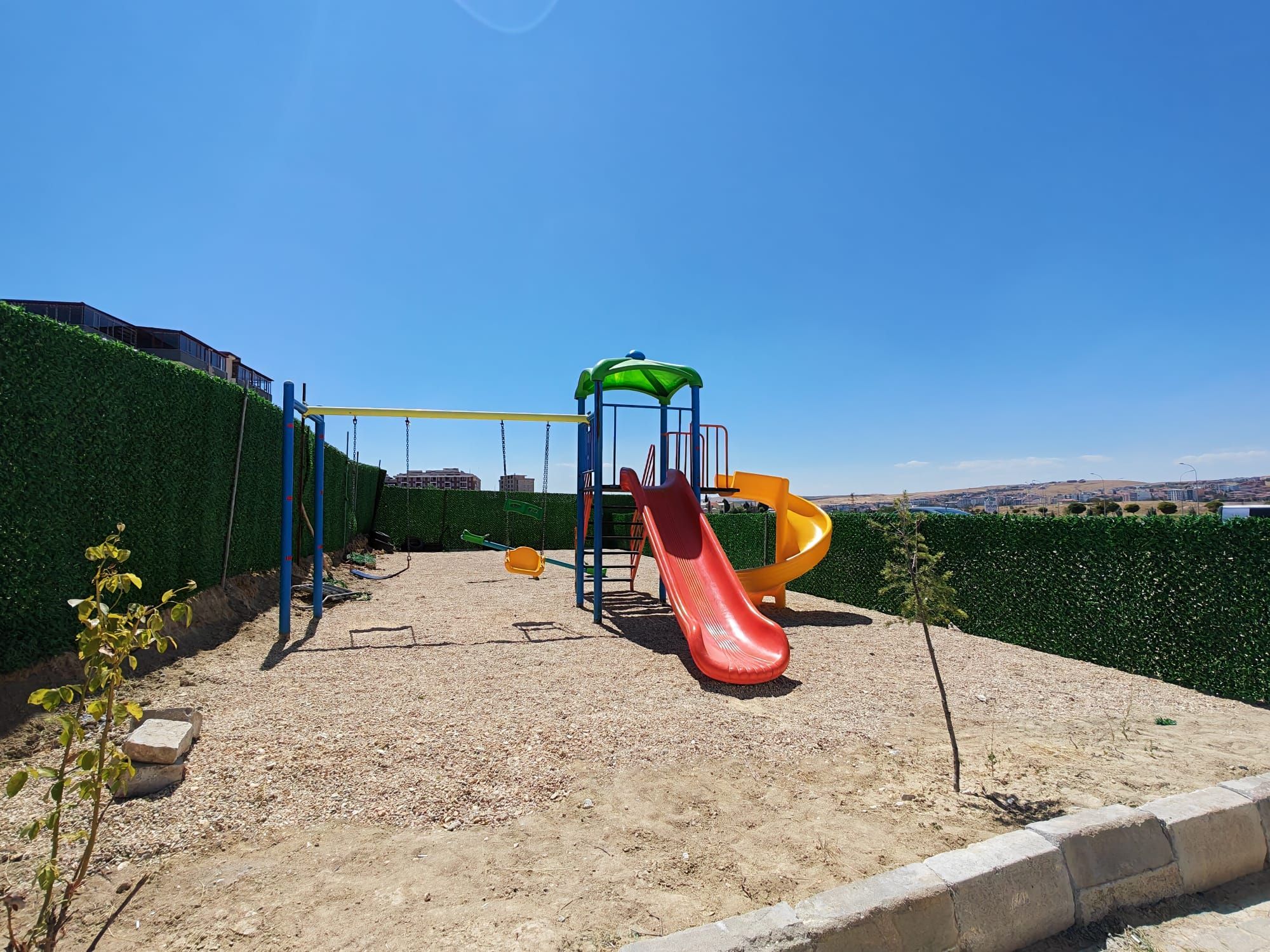 Une aire de jeux avec un toboggan et des structures d’escalade, entourée d’une haie et sous un ciel dégagé.