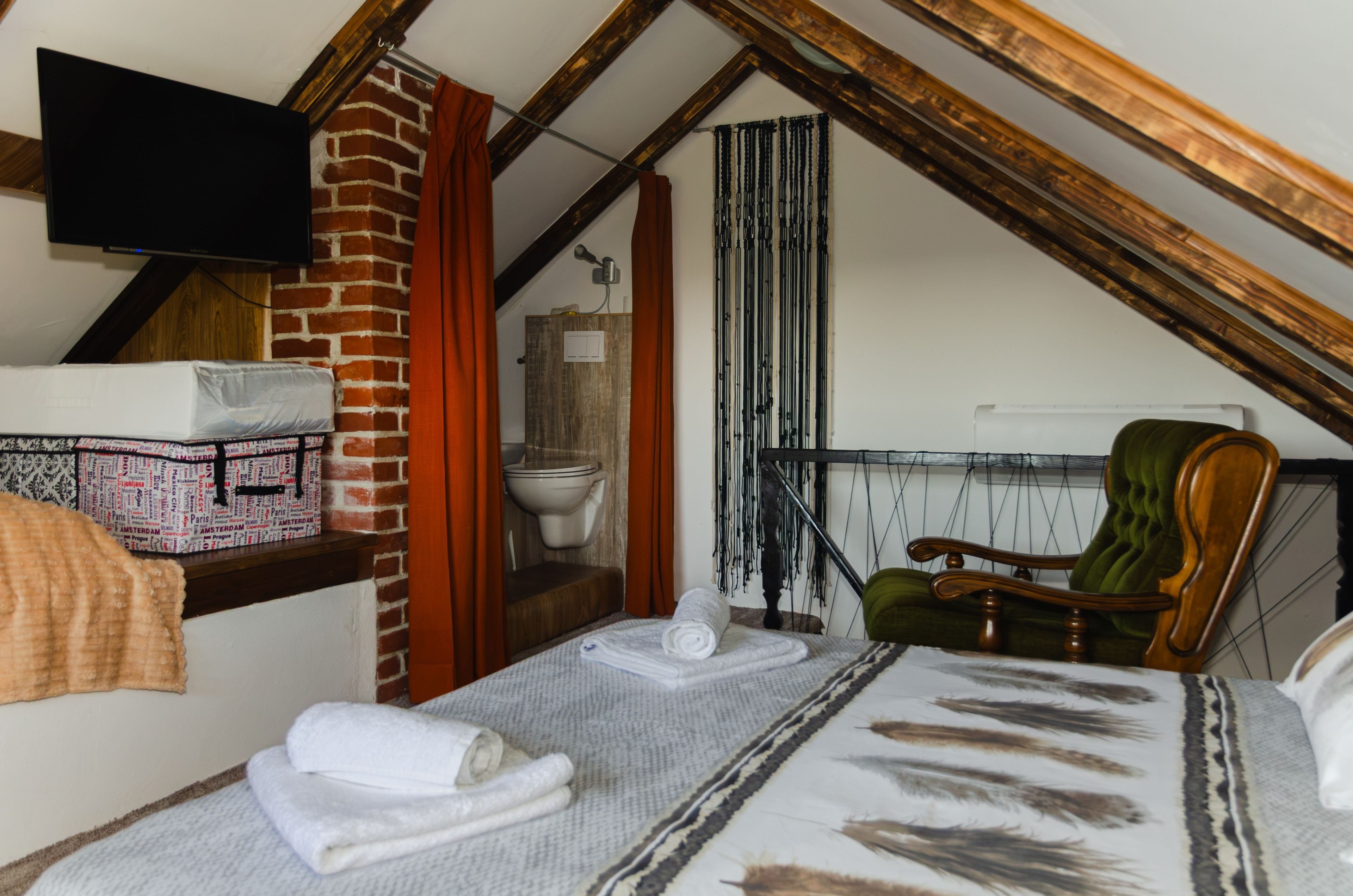 A small, cozy room with a sloped ceiling, a bed with a patterned blanket, a wooden chair, a television mounted on the wall, and a brick wall.