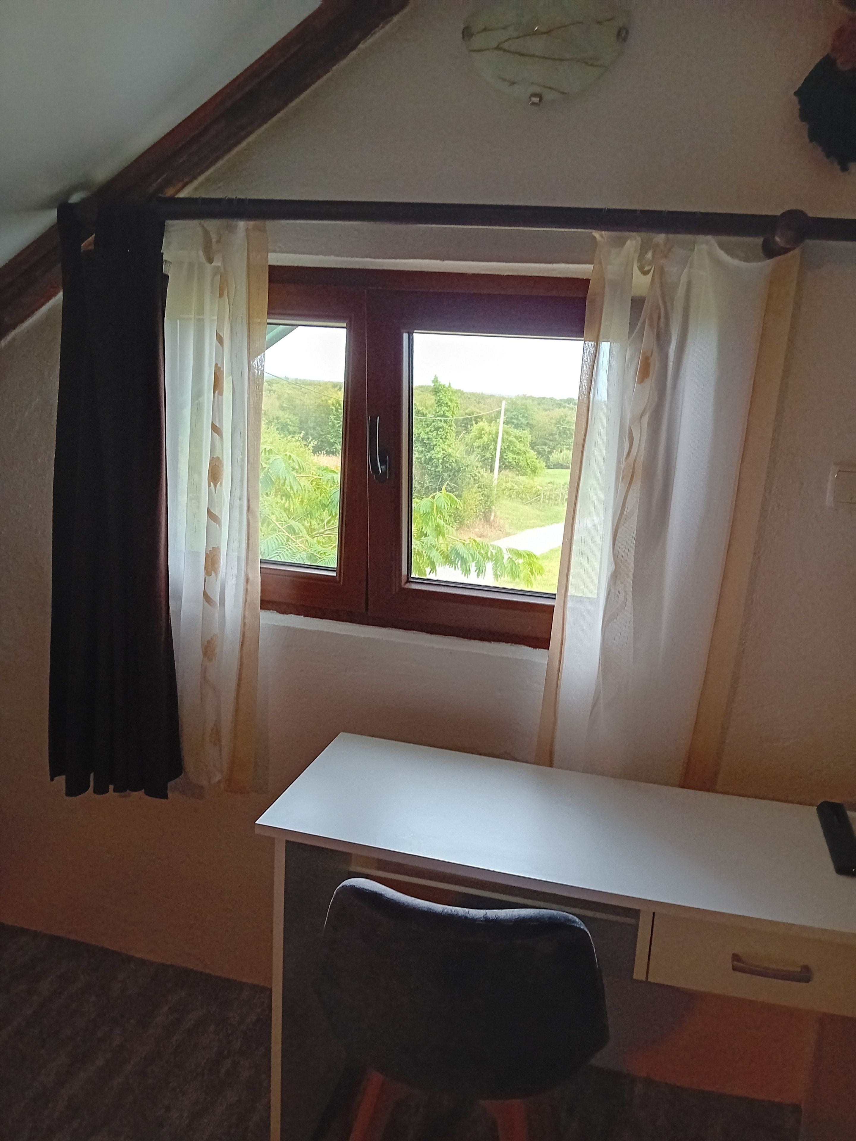 A room with a desk, a chair, a window with curtains, and a view of greenery outside.