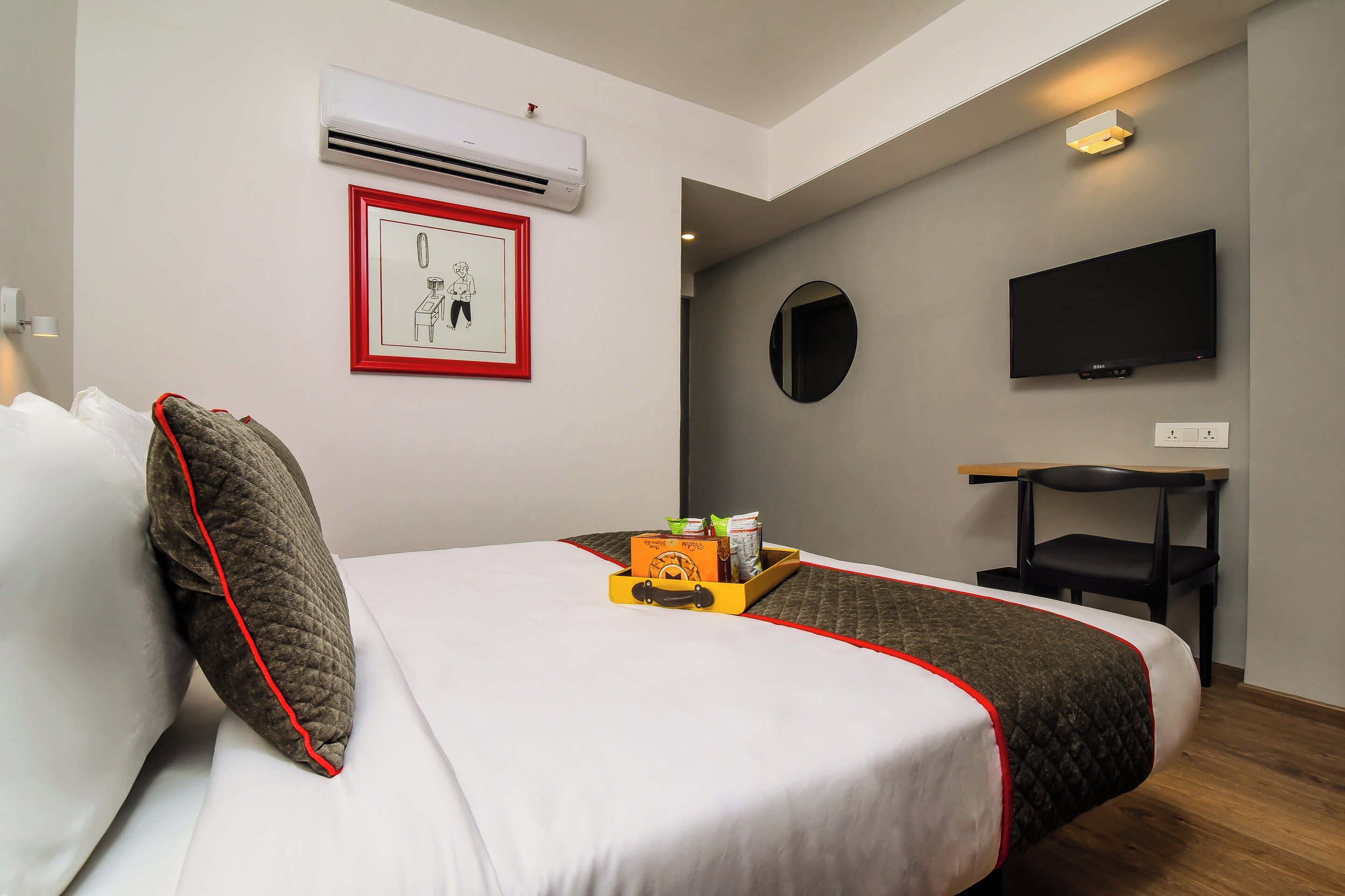 A hotel room with a bed, a flat-screen TV, an air conditioning unit, a framed picture, and a small table with a tray of items.