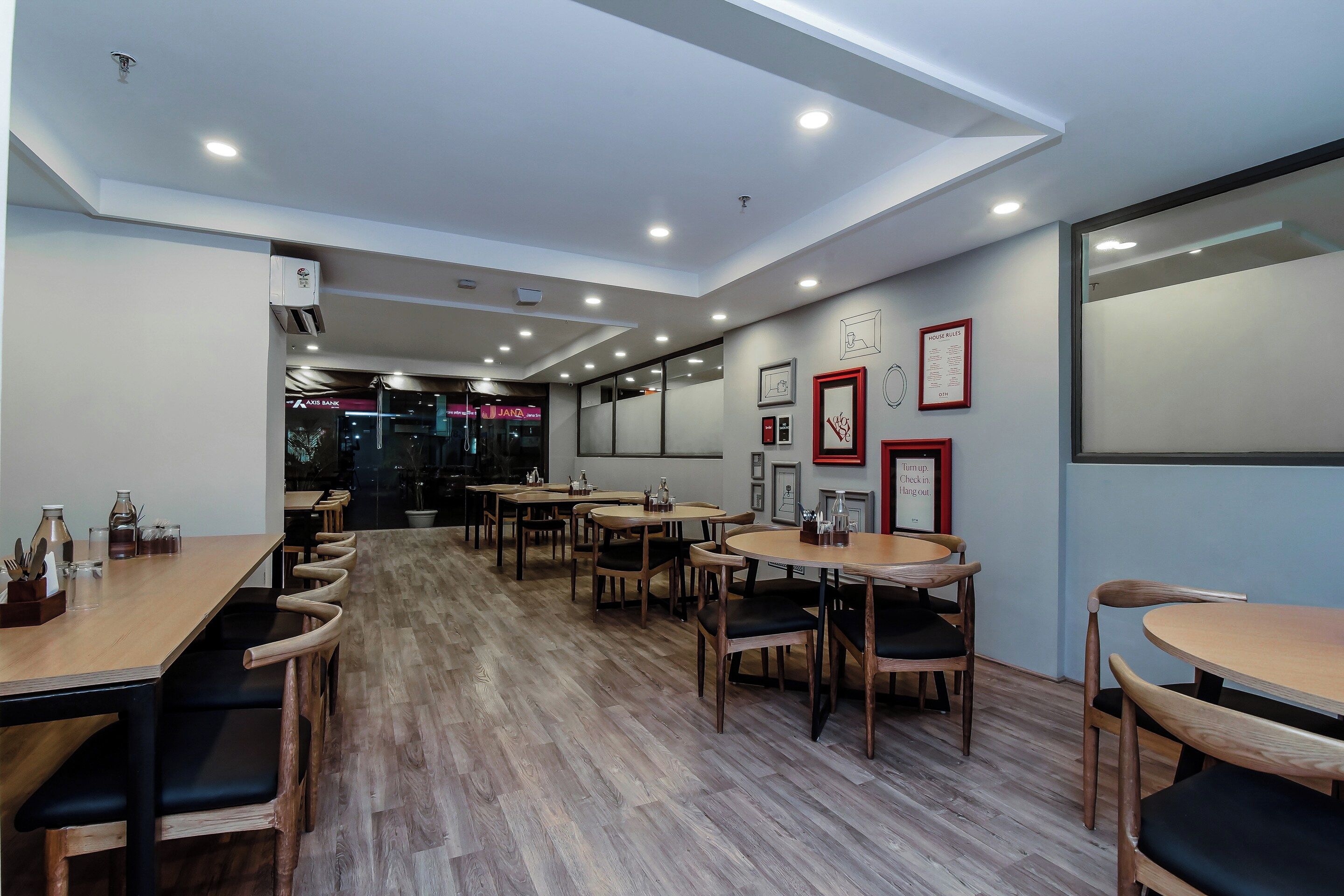 L’intérieur d’un restaurant moderne, avec des tables et des chaises en bois, un éclairage encastré au plafond et, au fond, une vue sur une banque et un magasin.