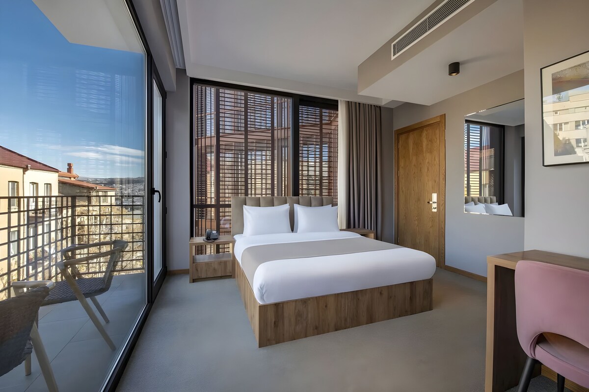 Ein modernes Hotelzimmer mit einem großen Bett, einem Schreibtisch, einem Stuhl und Blick auf die Stadt durch große Fenster.