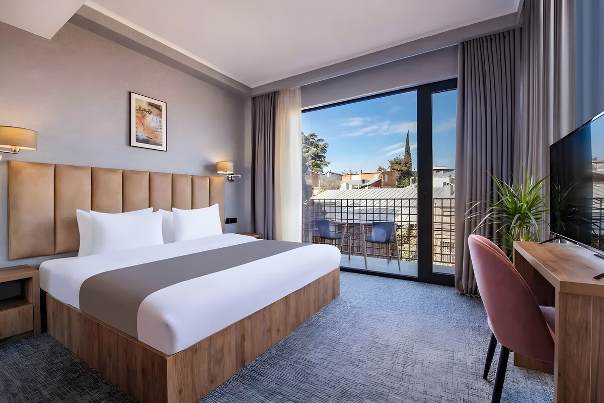 Ein Hotelzimmer mit einem großen Bett, einem Schreibtisch mit Stuhl, einem Fernseher und einem Balkon mit Stadtblick.