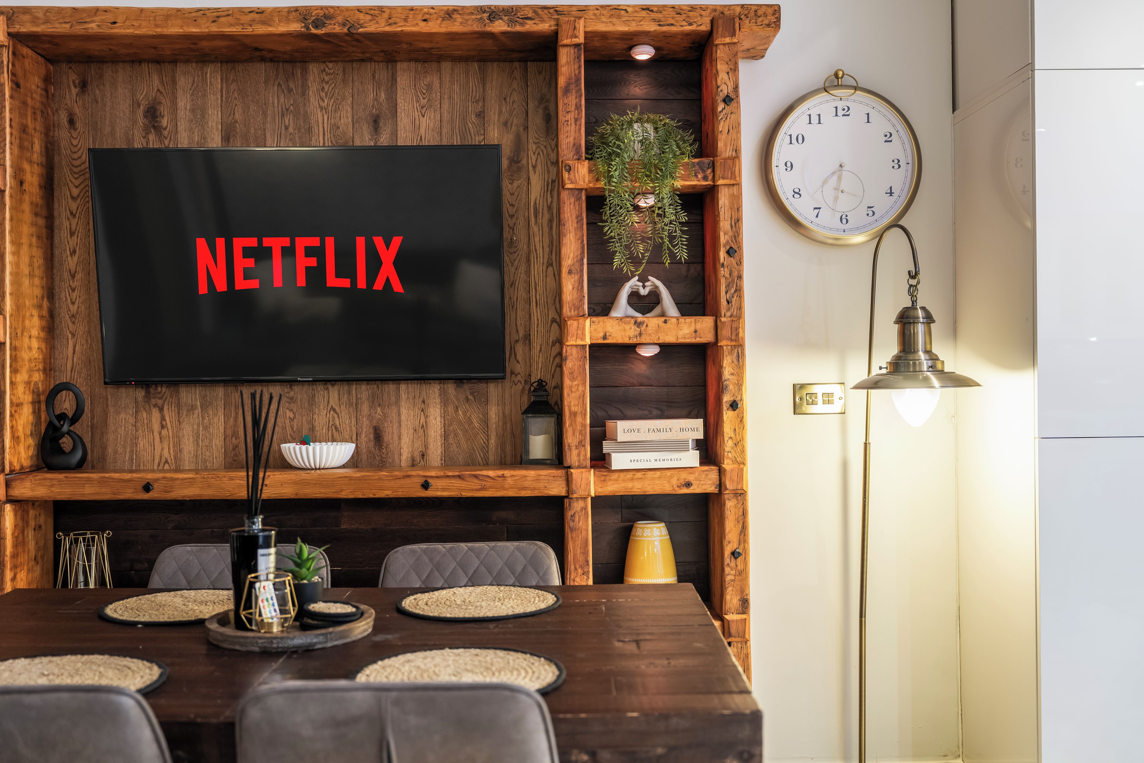 A wooden wall with a flat-screen TV displaying 'NETFLIX' in red letters, a clock, and a shelf with books and decorative items.