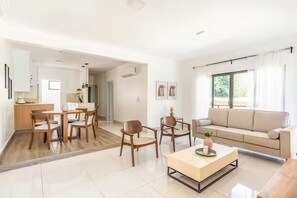 A modern living room with a sofa, coffee table, and chairs. There is a dining area with a table and chairs. The room has a large window with curtains. - Nazar Suites - Unit 2A (Santiago de los Caballeros)