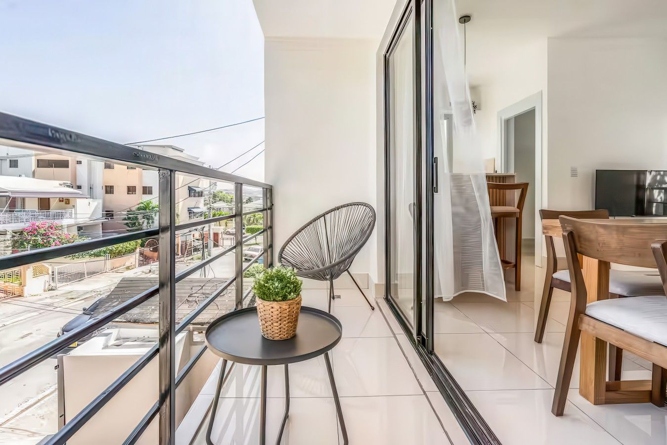 Un balcon donnant sur des immeubles, une petite table avec une plante, une chaise et un coin repas avec une table et des chaises.