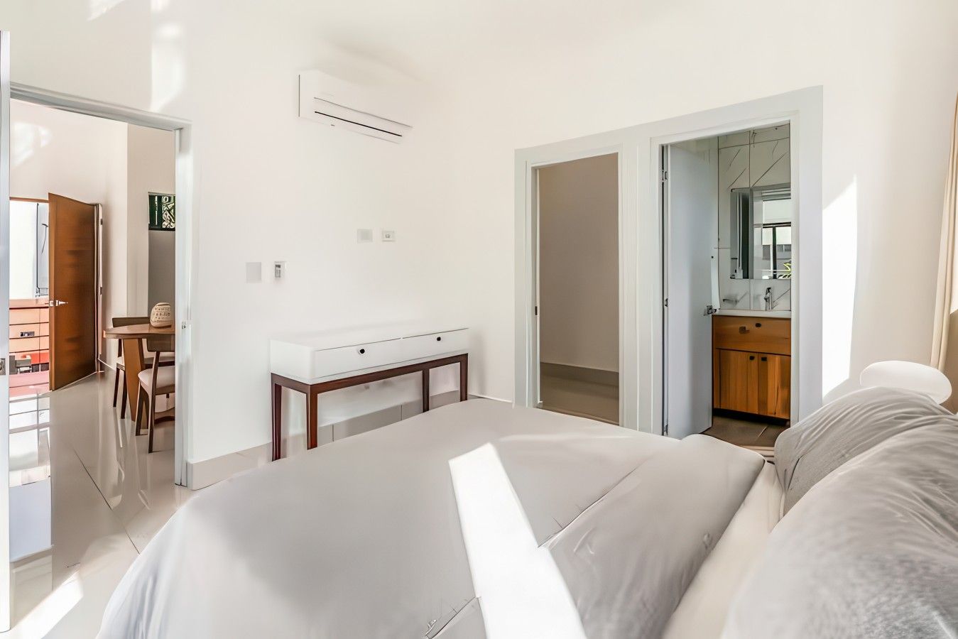 Ein Schlafzimmer mit Bett, Nachttisch und Kleiderschrank. Es gibt ein Badezimmer mit Spiegel und Waschbecken. Der Raum hat weiße Wände und eine Tür zu einem weiteren Zimmer.