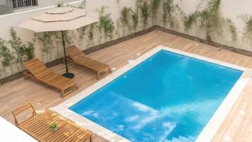 Ein rechteckiger Pool mit einer umlaufenden Holzterrasse, ausgestattet mit Liegestühlen und einem Tisch.