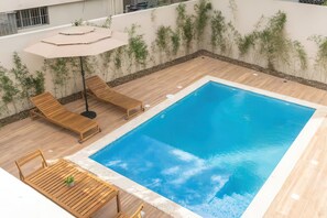 A rectangular swimming pool with a wooden deck surrounding it, equipped with lounge chairs and a table. - Nazar Suites - Entire 8-Bedroom Estate with Private Pool (Santiago de los Caballeros)