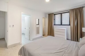 A bedroom with a large bed, a bedside table, a window with curtains, and a door leading to another room. - Nazar Suites - Unit 1A   (Santiago de los Caballeros)
