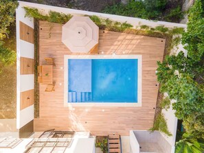 Een luchtendicht genomen beeld van een modern buitenzwembad met houten terrassen, omringd door groen en een witte muur.