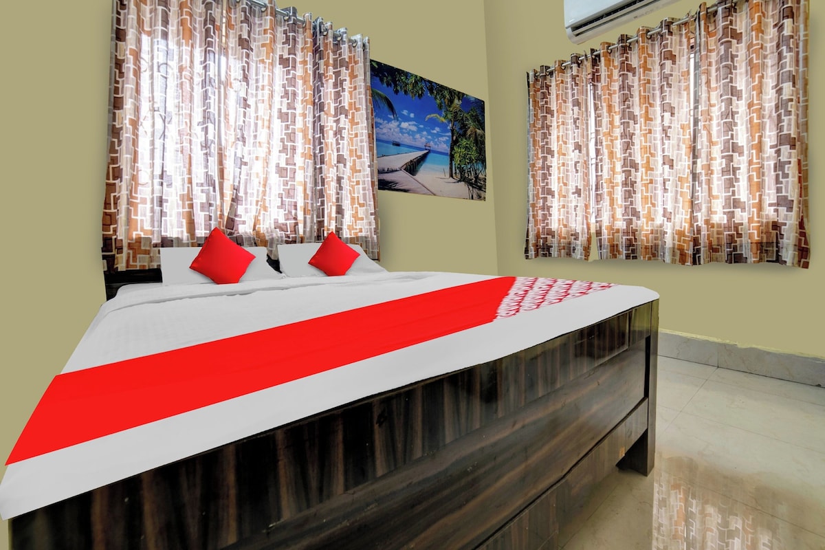 A hotel room with a bed, red pillows, patterned curtains, a painting of a beach scene, and an air conditioning unit.