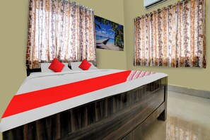 Habitación de hotel con cama, almohadas rojas, cortinas estampadas, un cuadro de una playa y un equipo de aire acondicionado.