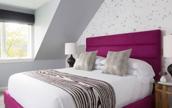 Un dormitorio con cama, almohadas, una mesita de noche, una lámpara y una ventana.