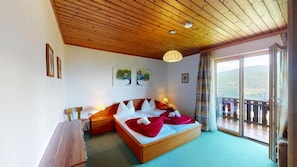 Ein Schlafzimmer mit Holzboden, einem großen Bett mit roter Bettwäsche, einem Nachttisch, einem Schreibtisch und Blick ins Freie durch eine Schiebetür.