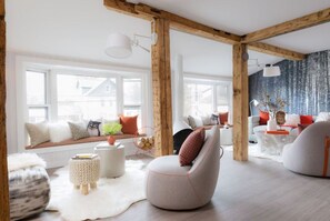 Una sala de estar moderna con vigas de madera a la vista, un amplio banco junto a la ventana con cojines, un sillón blanco redondo y una alfombra blanca.