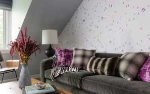 Una sala de estar acogedora con un sofá gris, cojines morados y marrones, un jarrón con flores y una lámpara.