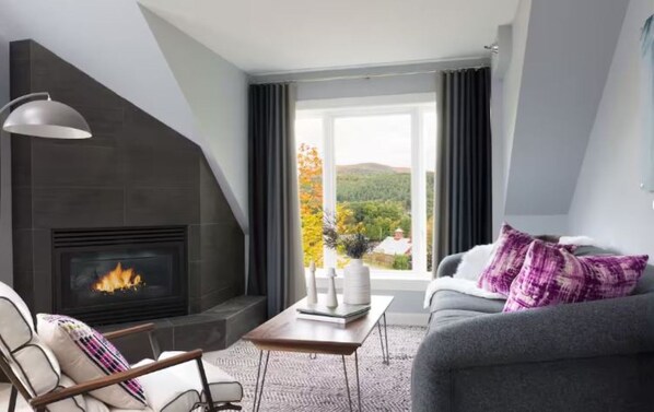 Una sala de estar moderna con chimenea, un sofá, una mesa de centro y vistas a un área boscosa.