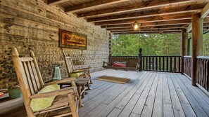 A wooden deck with a bench and chairs, a hanging light fixture, and a view of greenery. - Timberstone: Luxury 4BR Cabin, Pool table, Arcade, Hot Tub, & Resort Pool (Sevierville)