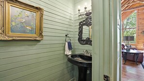 Casa de banho com parede verde, quadro com pássaros, pia, espelho e porta-toalhas.