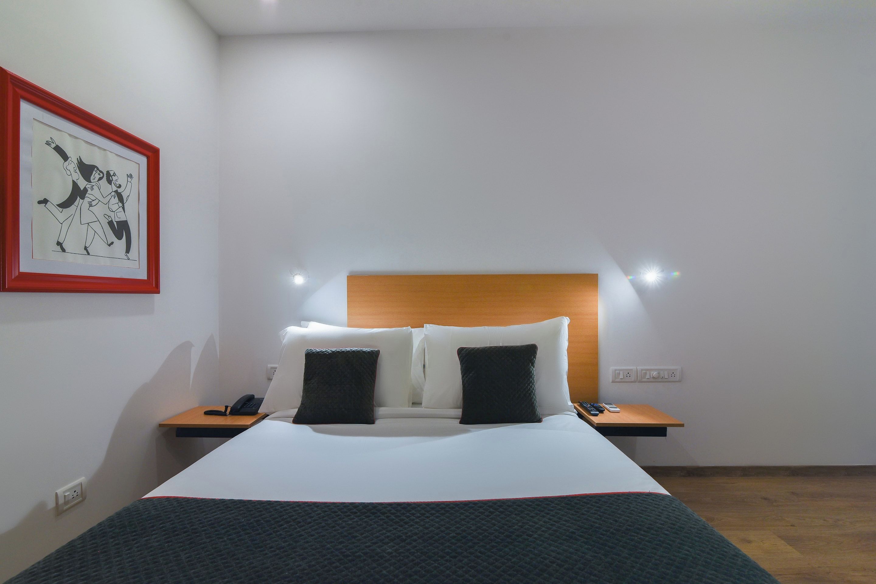 Una habitación de hotel con una cama, dos mesitas de noche, una lámpara de pared e imagen en un marco.