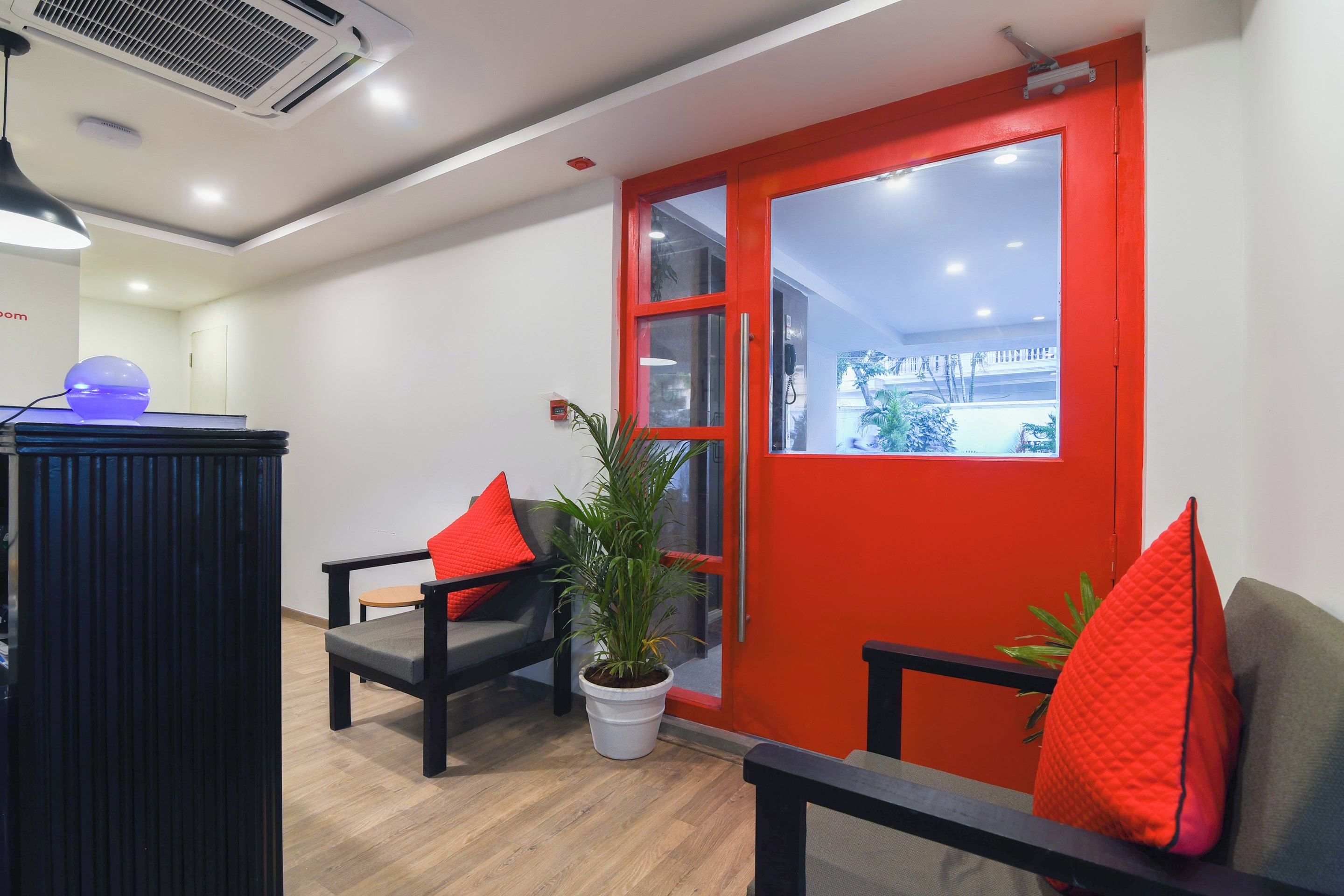 A reception area with a red door, two black chairs with red cushions, a potted plant, and a desk with a blue object on it.