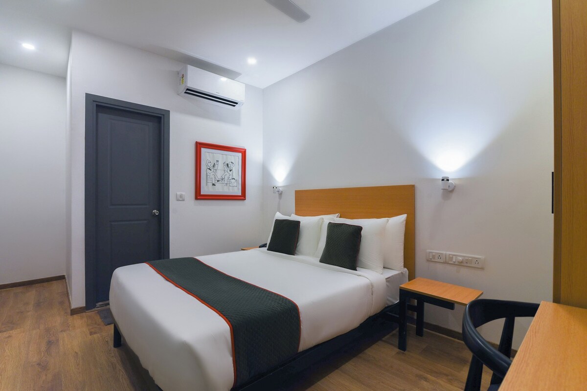 Quarto de hotel com cama, escrivaninha, cadeira, ar condicionado e quadro na parede.