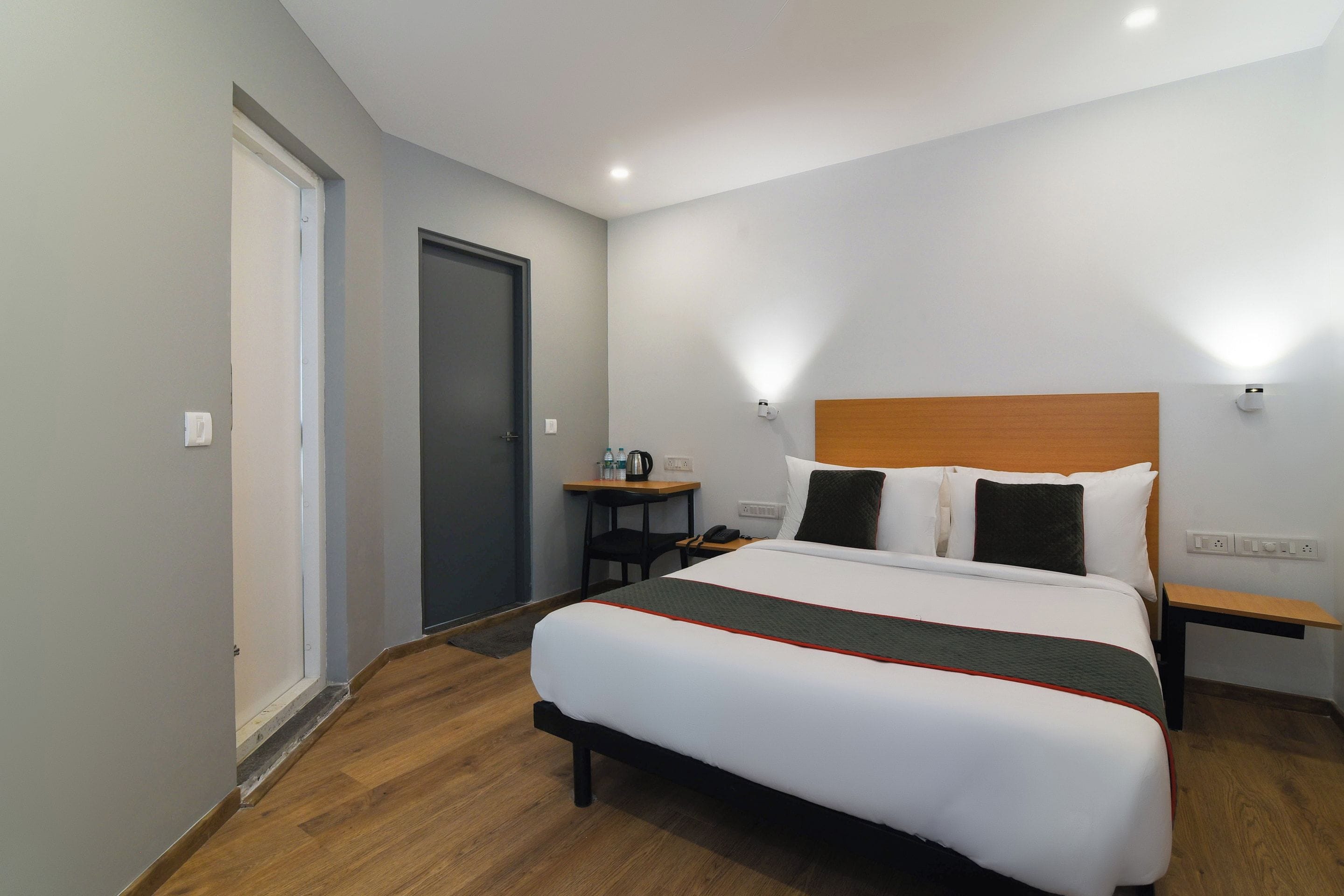 Habitación de hotel con cama, mesitas de noche, escritorio, silla, puerta y aplique de pared.