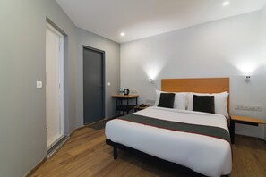 A hotel room with a bed, bedside tables, a desk, a chair, a door, and a wall-mounted light fixture.