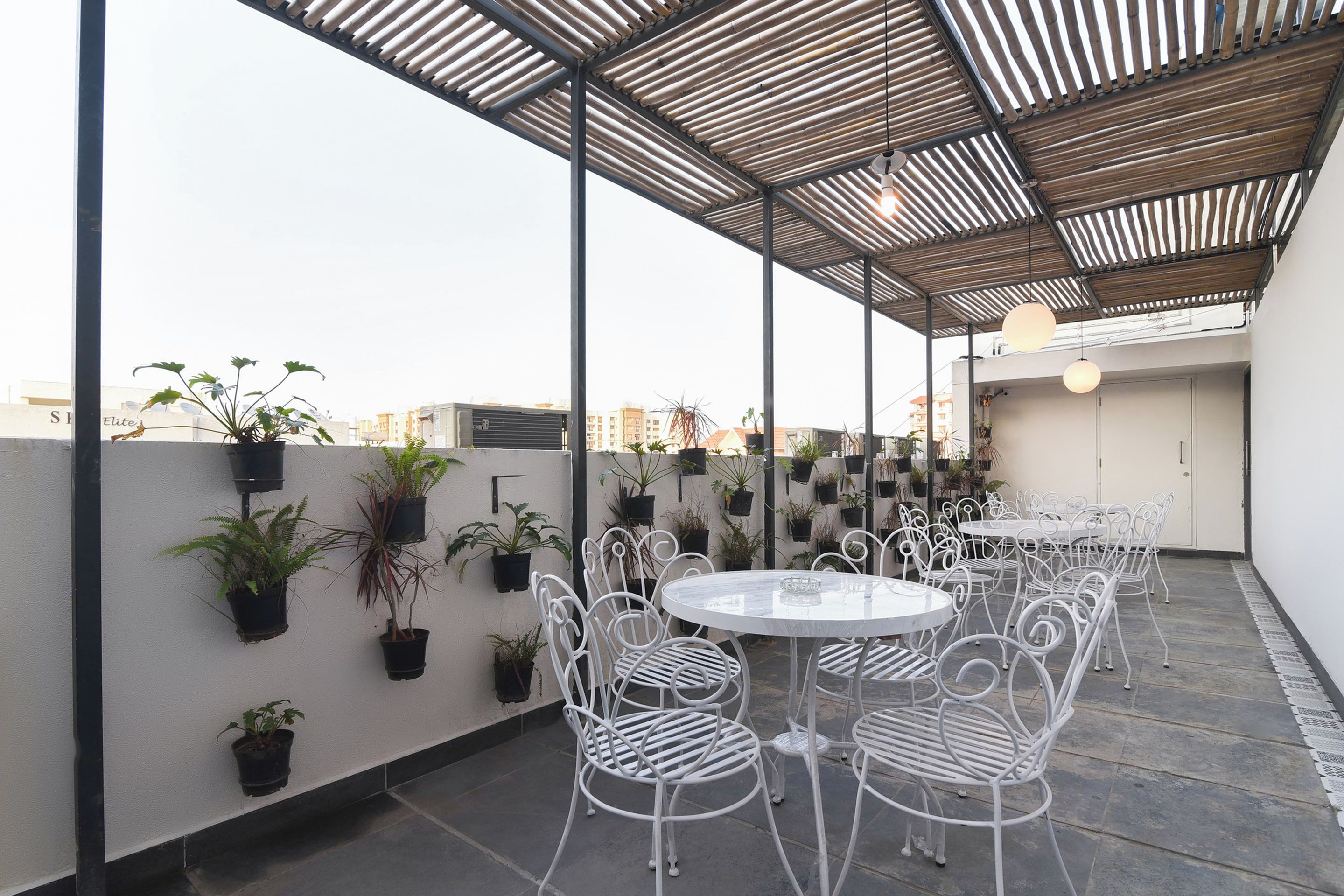 Una terraza en la azotea con mesas redondas y sillas de metal blanco, rodeada de plantas en macetas.