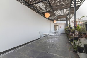 Um pátio externo com mesas e cadeiras de metal, piso de azulejos e plantas em vasos.