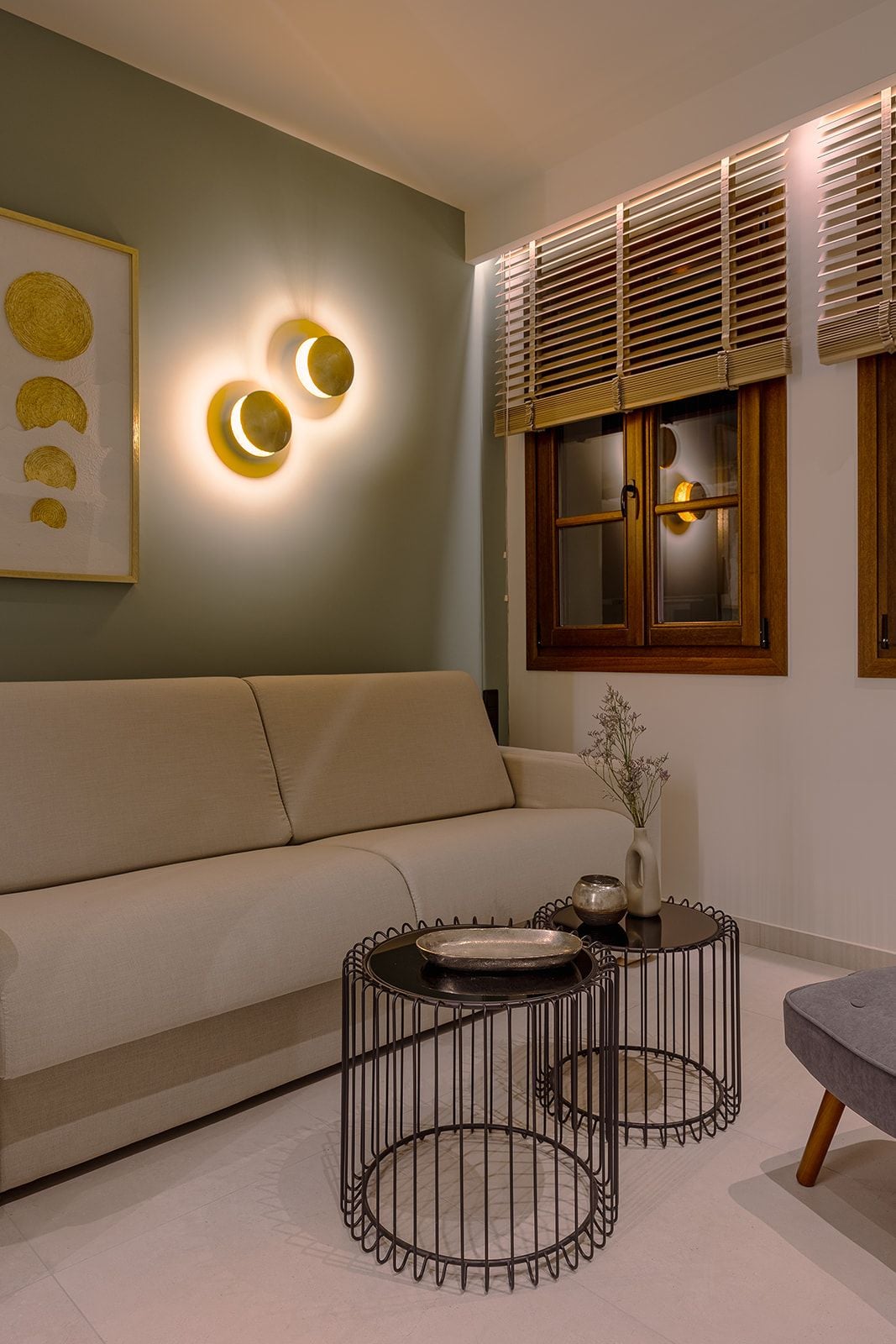 Una sala de estar moderna con un sofá, dos mesas redondas de metal, un ventanal con persianas, iluminación empotrada en la pared y una obra de arte enmarcada.
