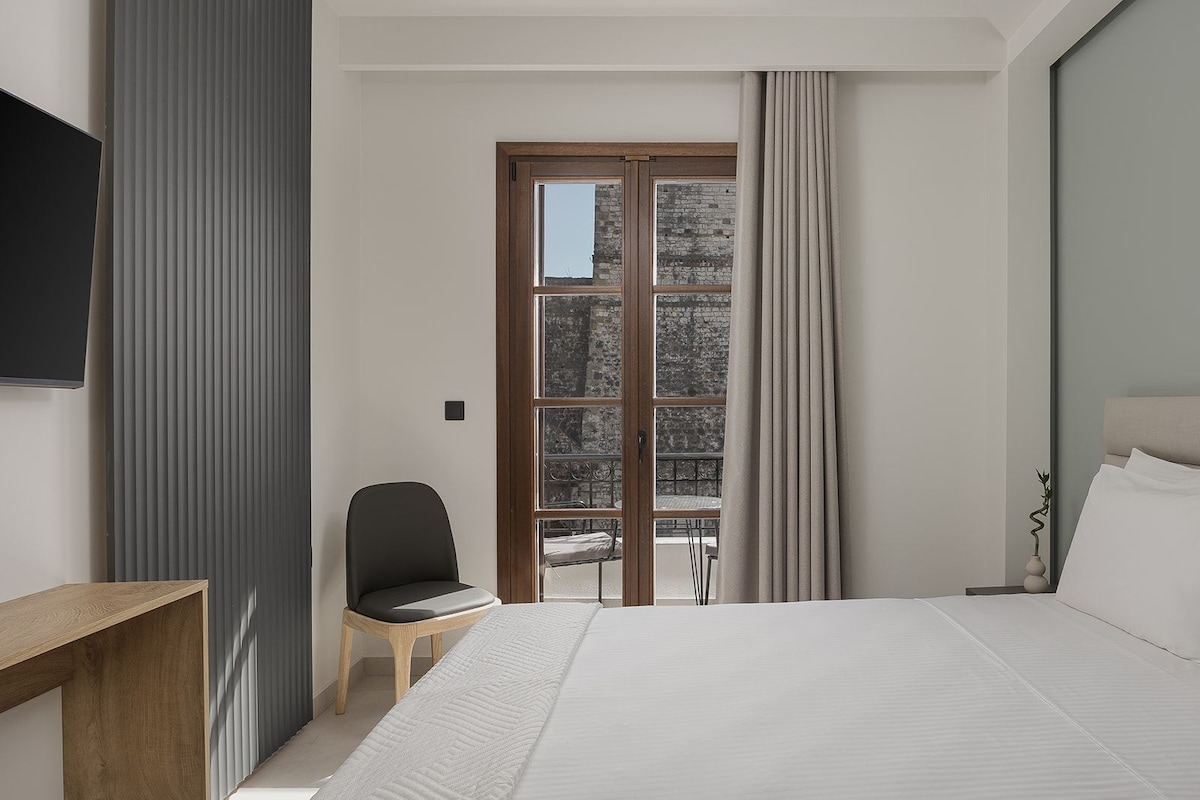 Habitación de hotel con una cama, una silla, una mesa de madera y un gran ventanal con cortinas.