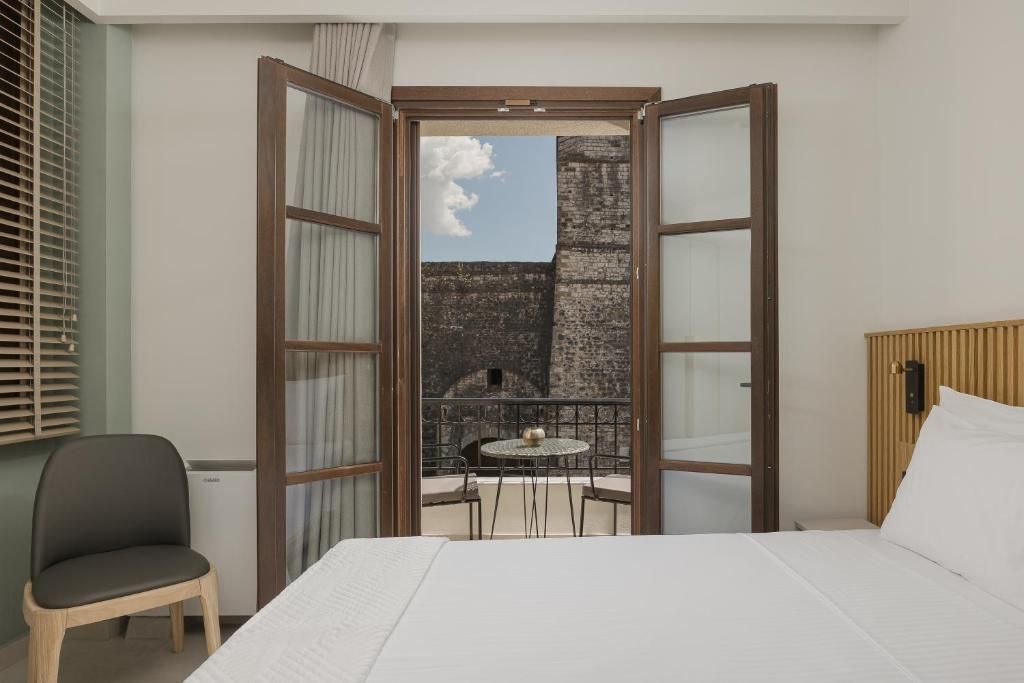 A hotel room with a bed, a chair, a table, and a view of an outdoor area with a brick wall and a clear sky.