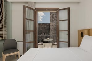 A hotel room with a bed, a chair, a table, and a view of an outdoor area with a brick wall and a clear sky. - Elysian Castle (Ioannina)