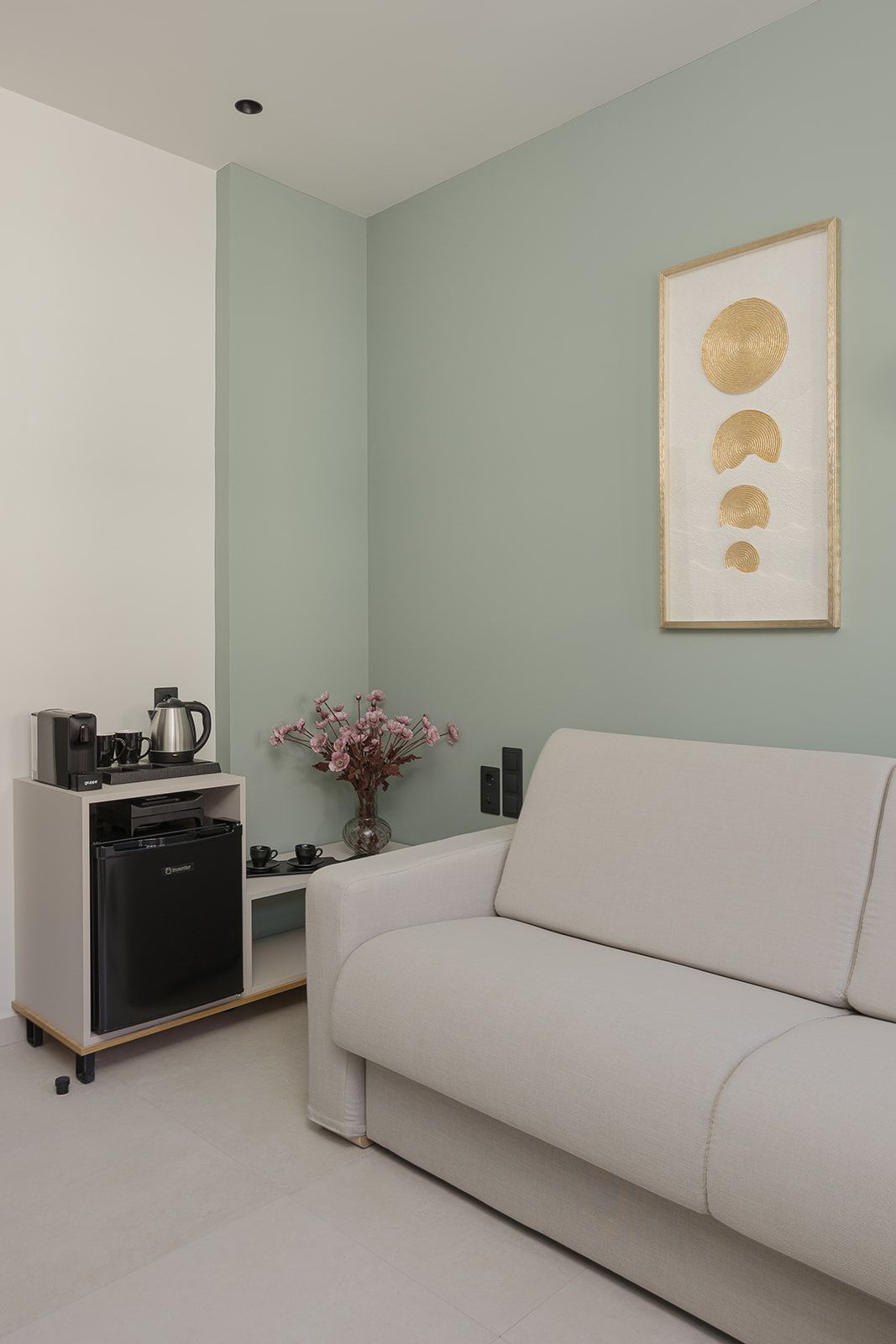 A modern living room with a sofa, a coffee machine, a small table with a vase of flowers, and a framed artwork on the wall.