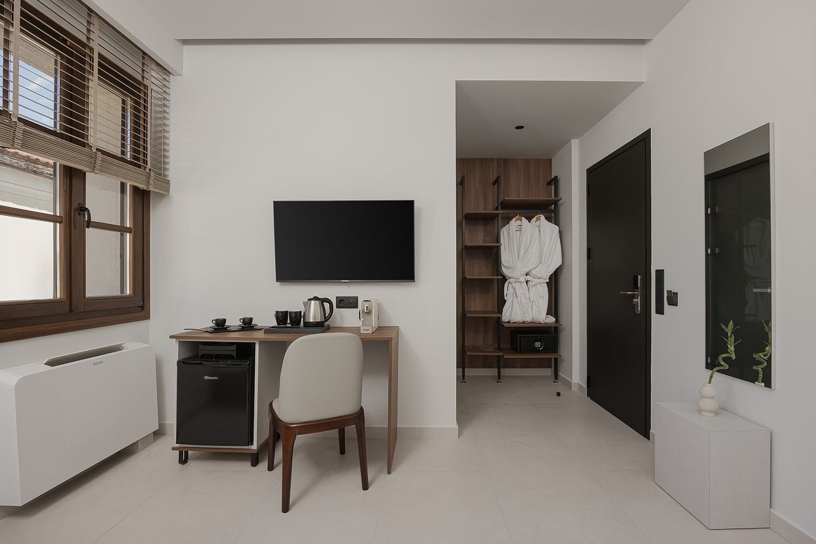 A modern hotel room with a kitchenette, a flat-screen TV, a wooden desk, a chair, a wardrobe, and a mirror.