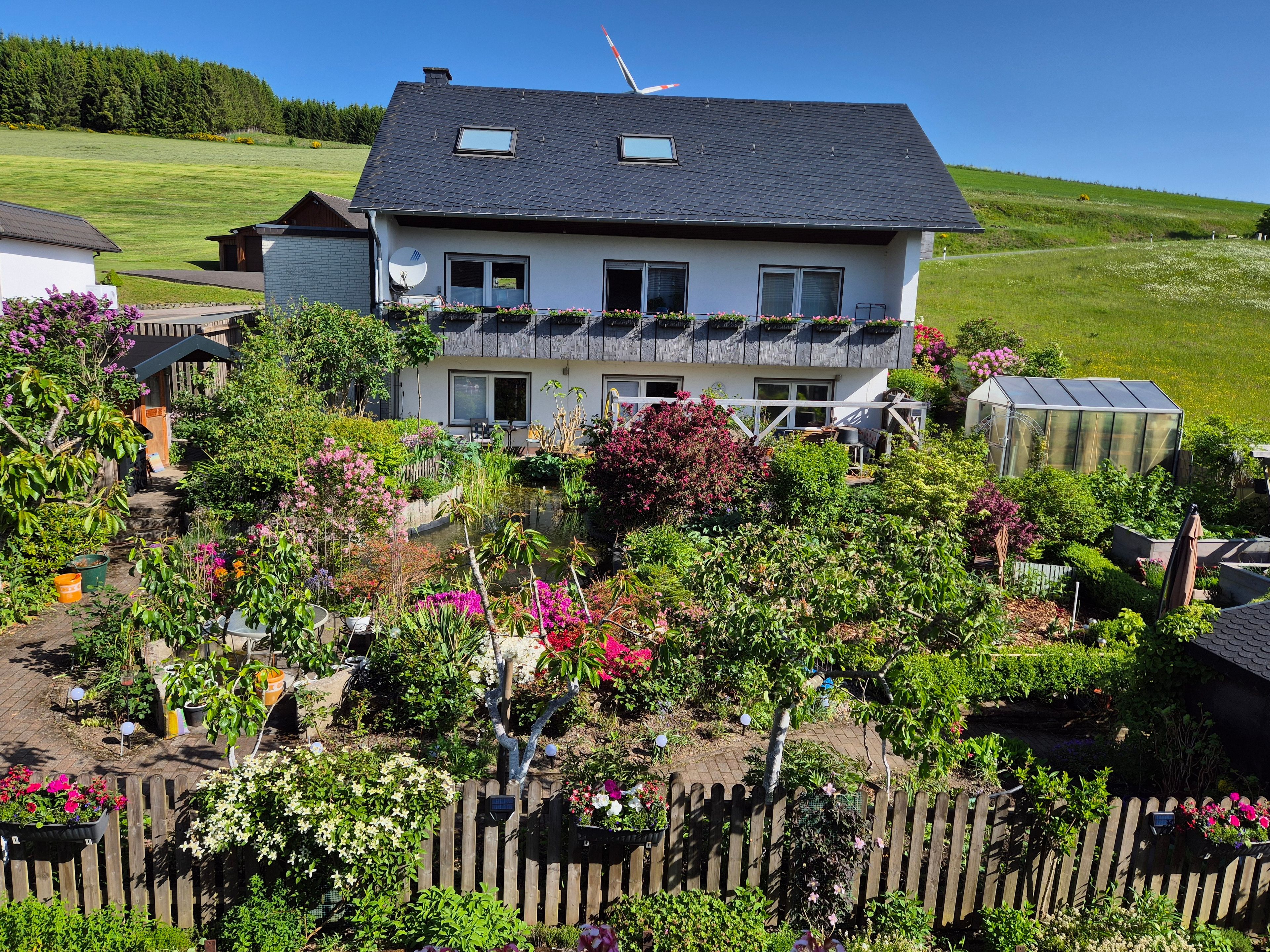 Une maison à deux étages avec un jardin, une serre et une clôture en bois.